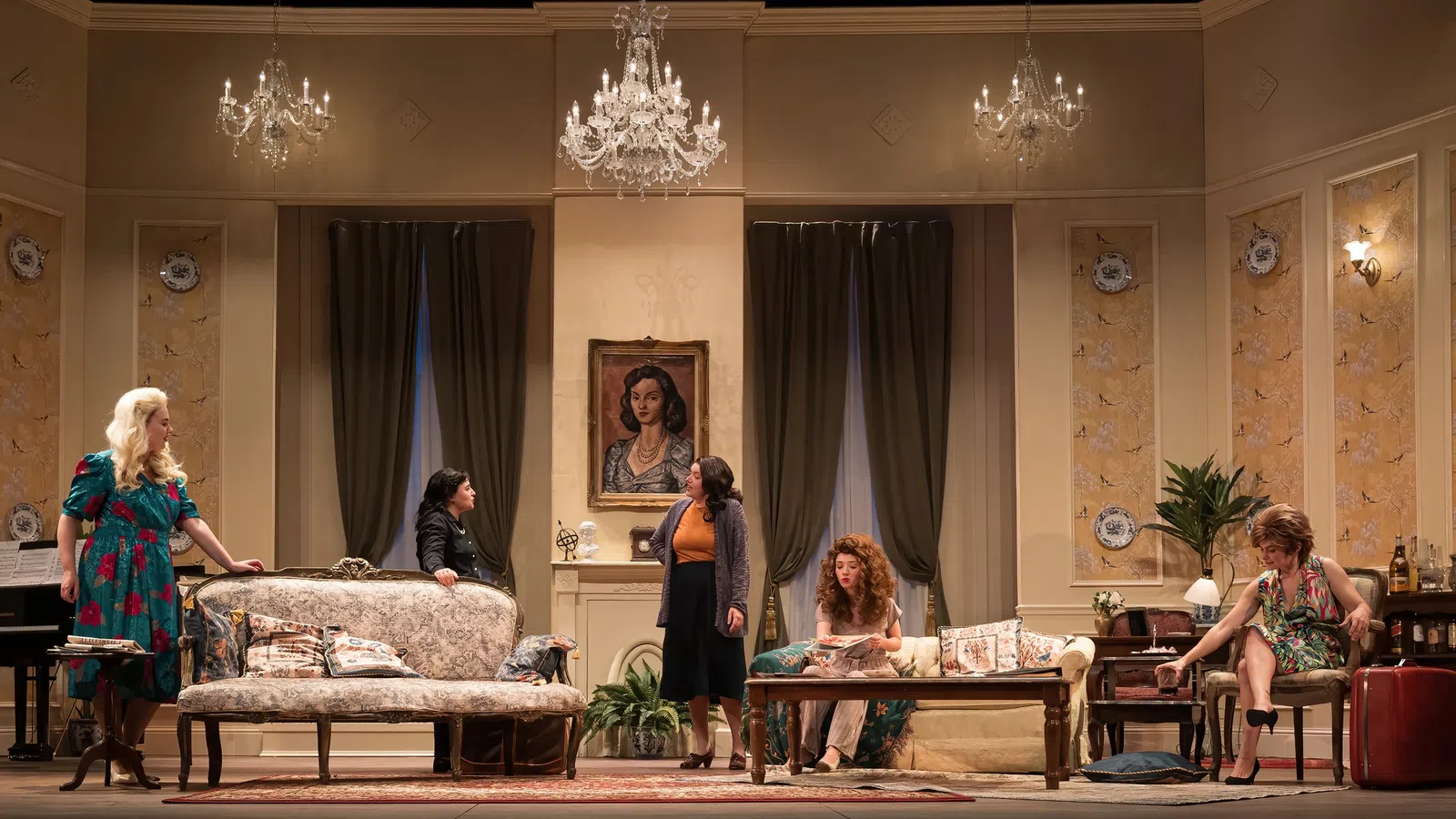 Theatrical stage scene with five women in vintage living room setting under chandeliers and art-adorned walls