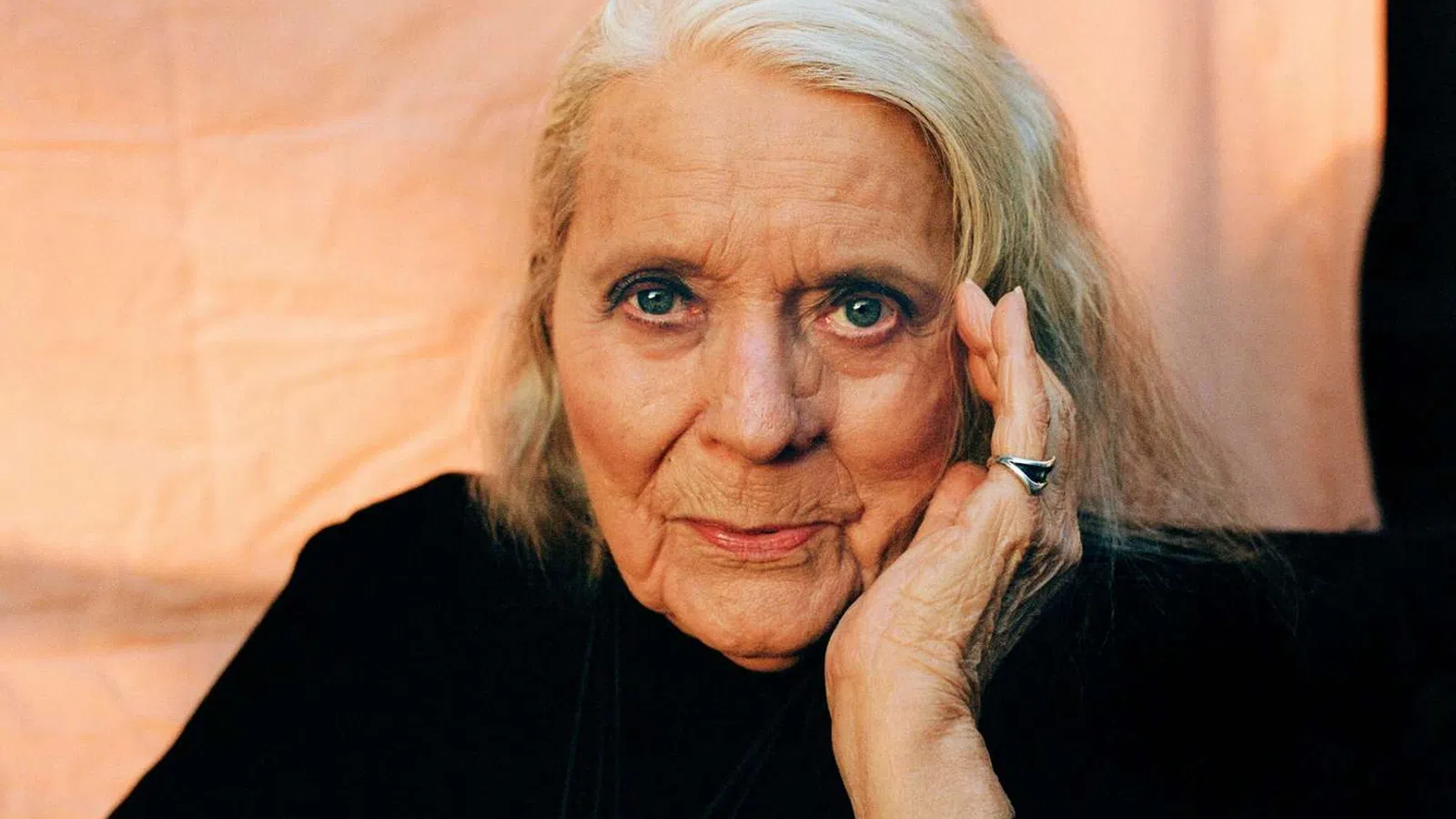 Elderly woman with silver hair, wearing black, resting her face on her hand against a soft peach background.