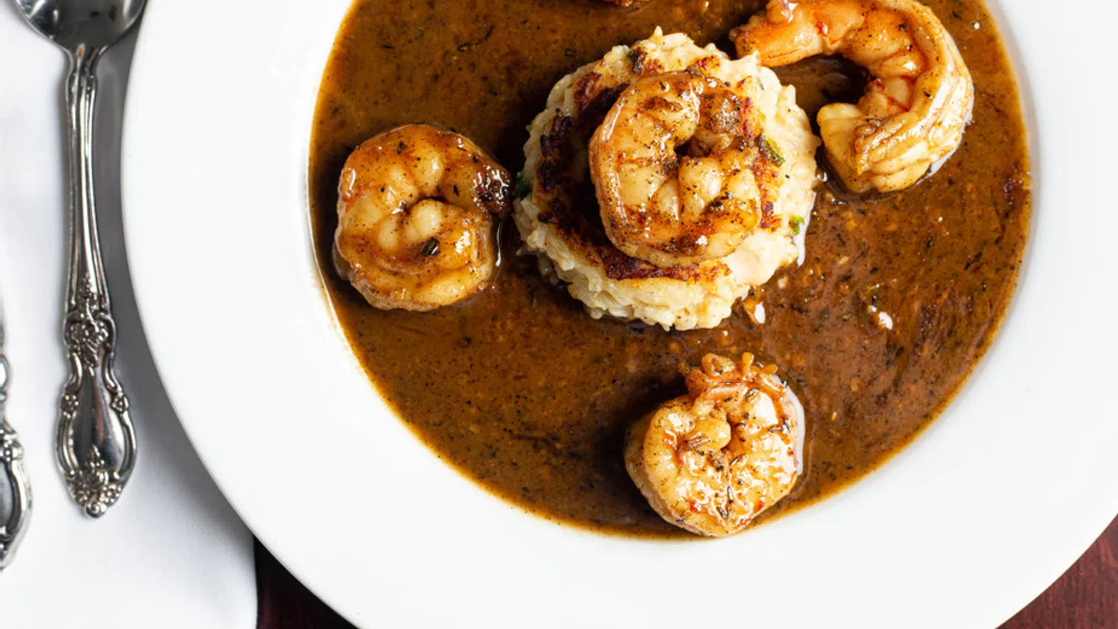 Spicy shrimp served over creamy grits with rich brown sauce on a white plate beside vintage silverware.
