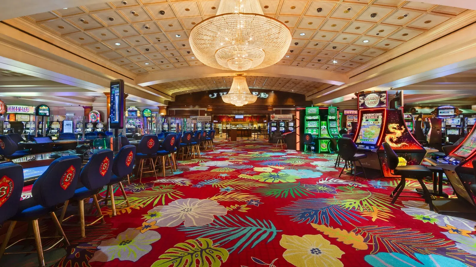 Colorful casino interior with floral carpet, gaming machines, blue chairs, and large chandeliers overhead