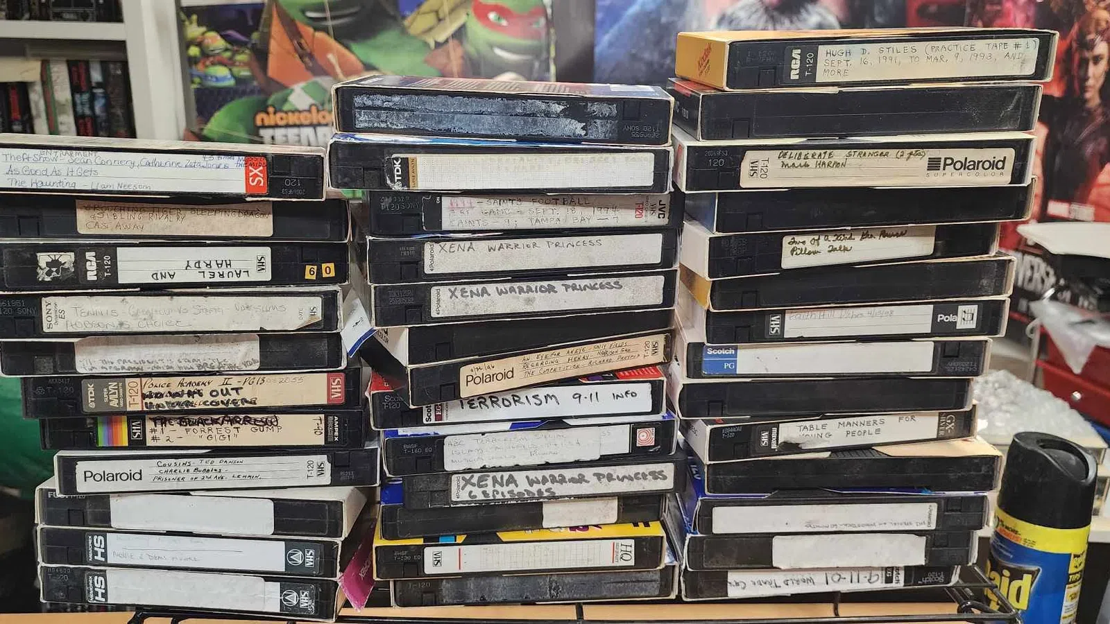 Stack of vintage VHS tapes with handwritten labels on a cluttered desk surrounded by posters and household items.
