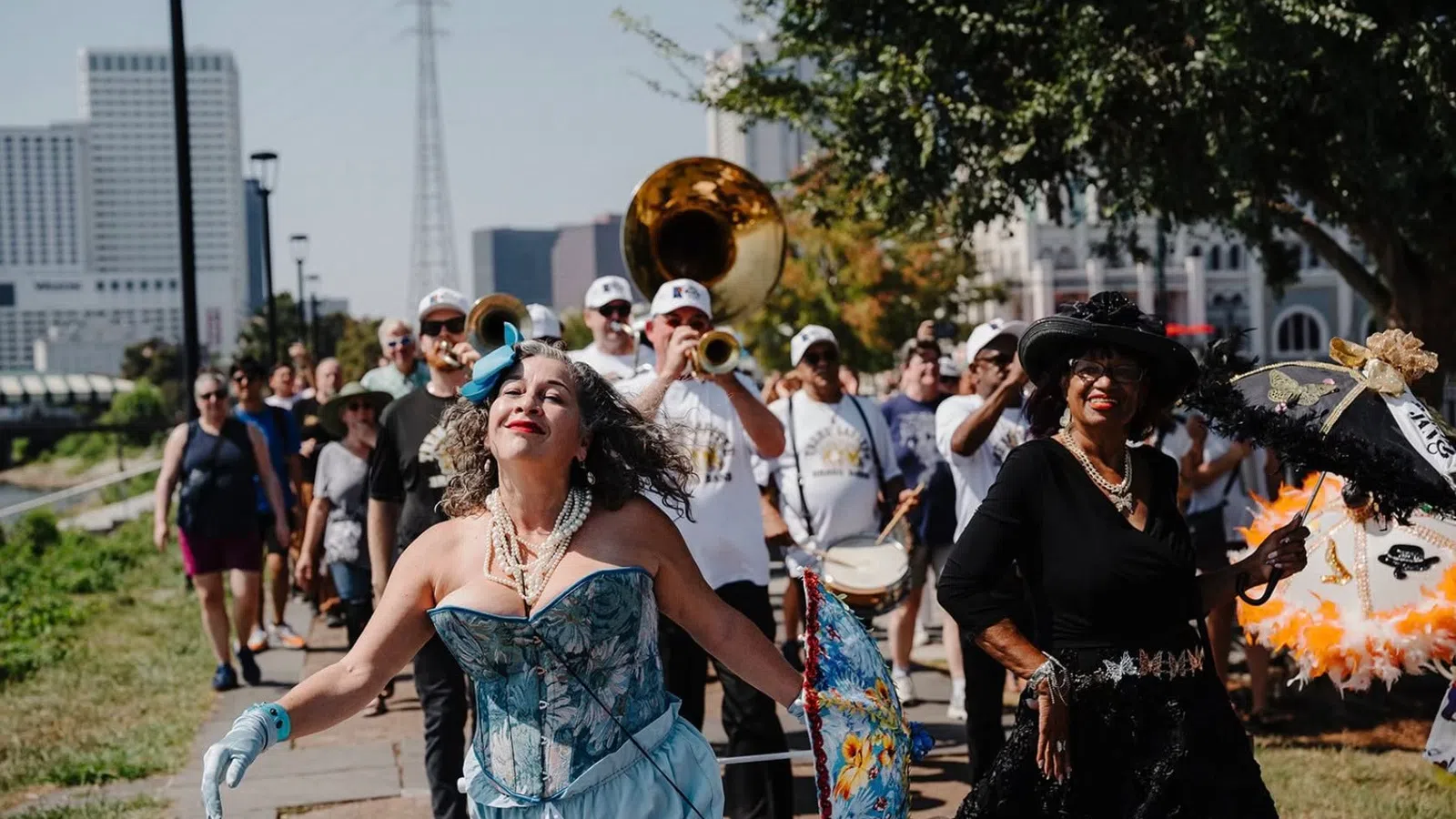 Woman in blue corset and gloves dances joyfully in front of a marching band and parade crowd on sunny day.
