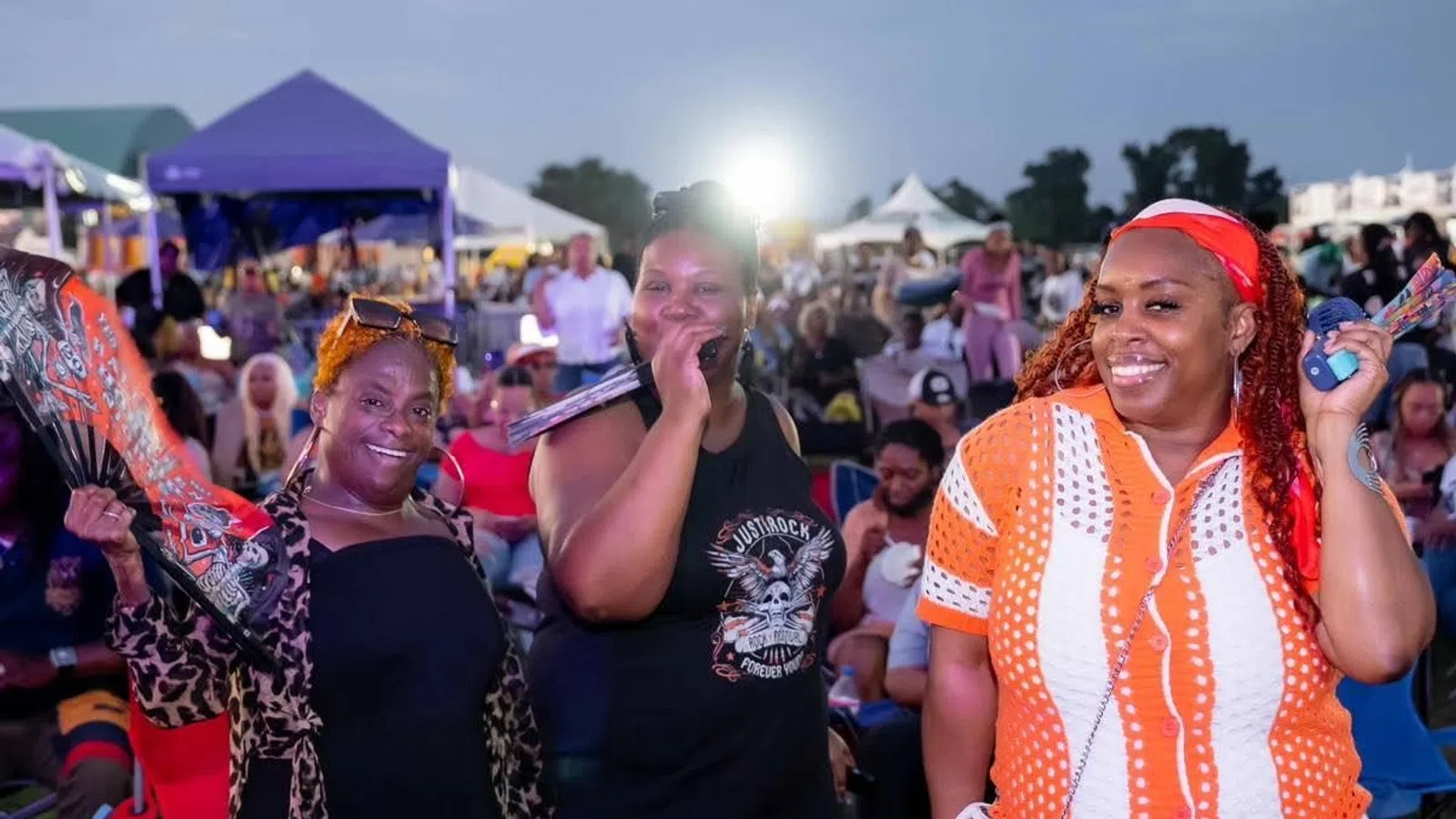 Three smiling women at an outdoor event with tents and crowd in the background during evening.
