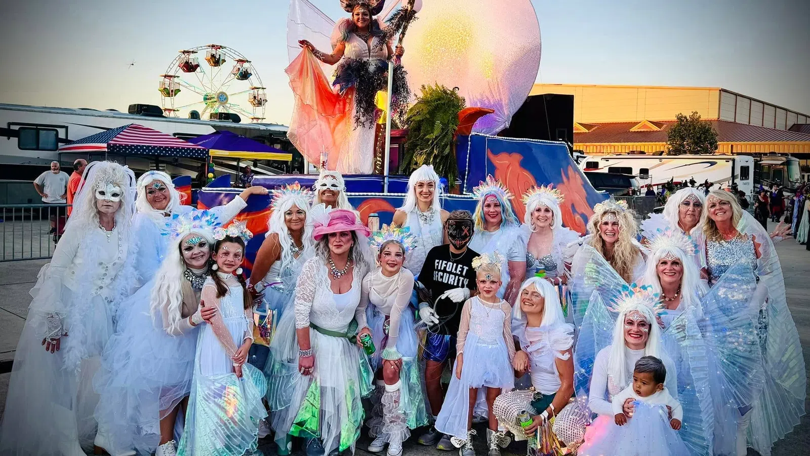 Group of people in white costumes and wings posing outdoors at a festival with a ferris wheel and colorful float.