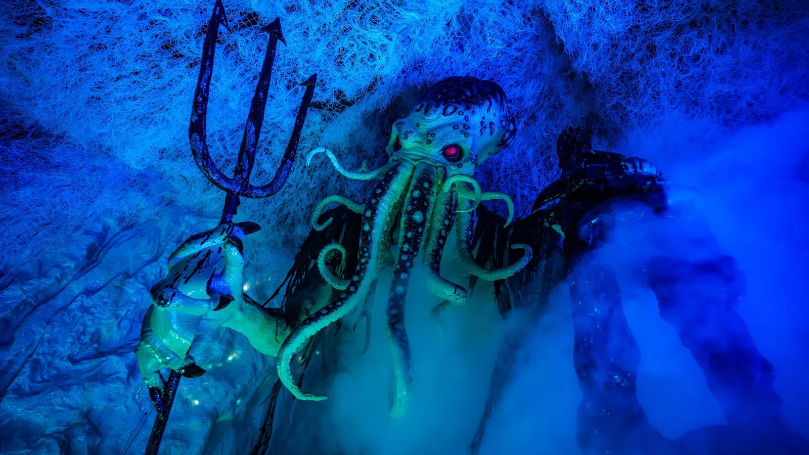Cthulhu figure with glowing red eyes holding a trident amidst blue fog and web-like textures underwater.
