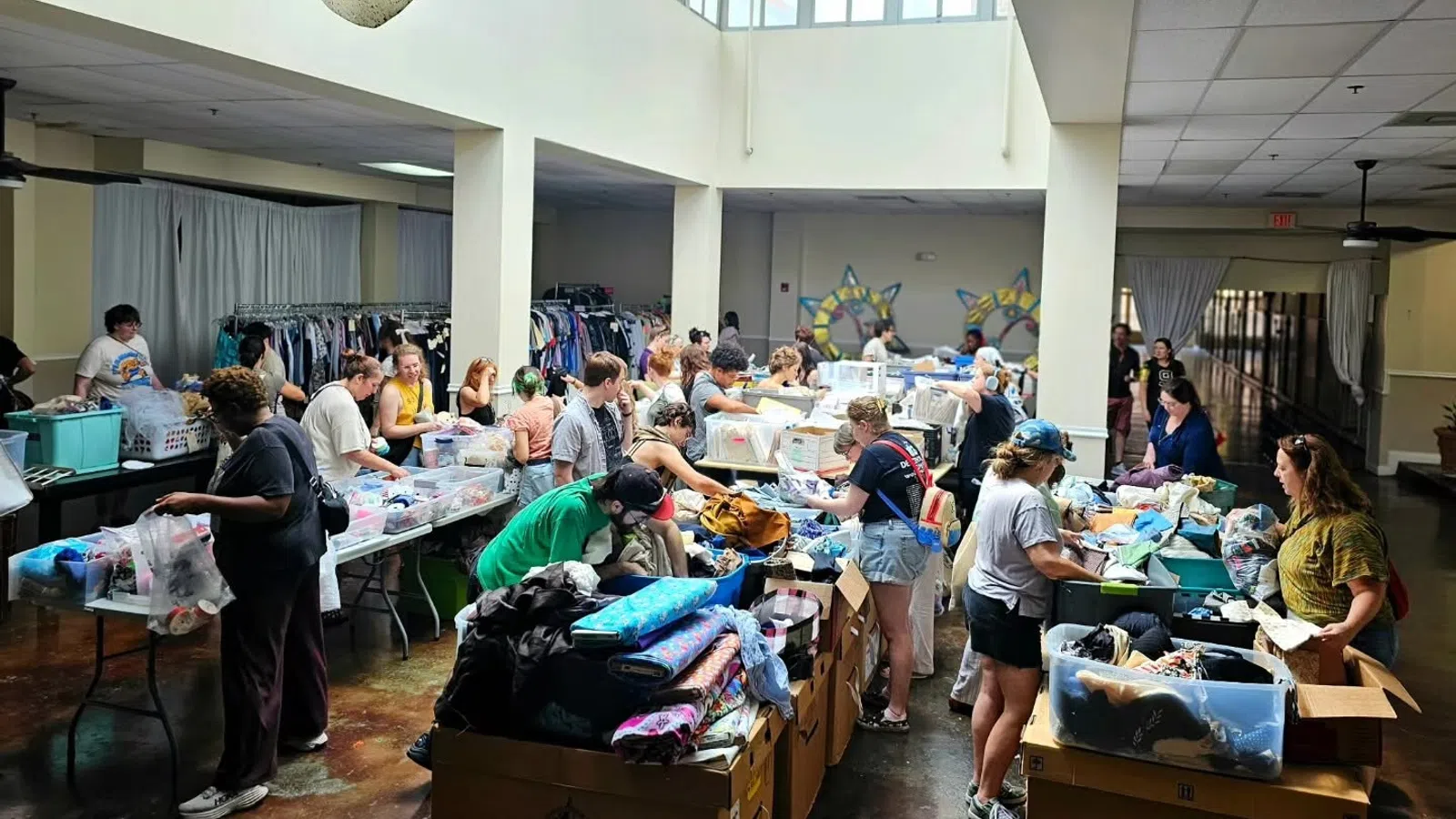 Volunteers sorting and organizing clothing donations in a large, well-lit community center with boxes and racks of clothes.