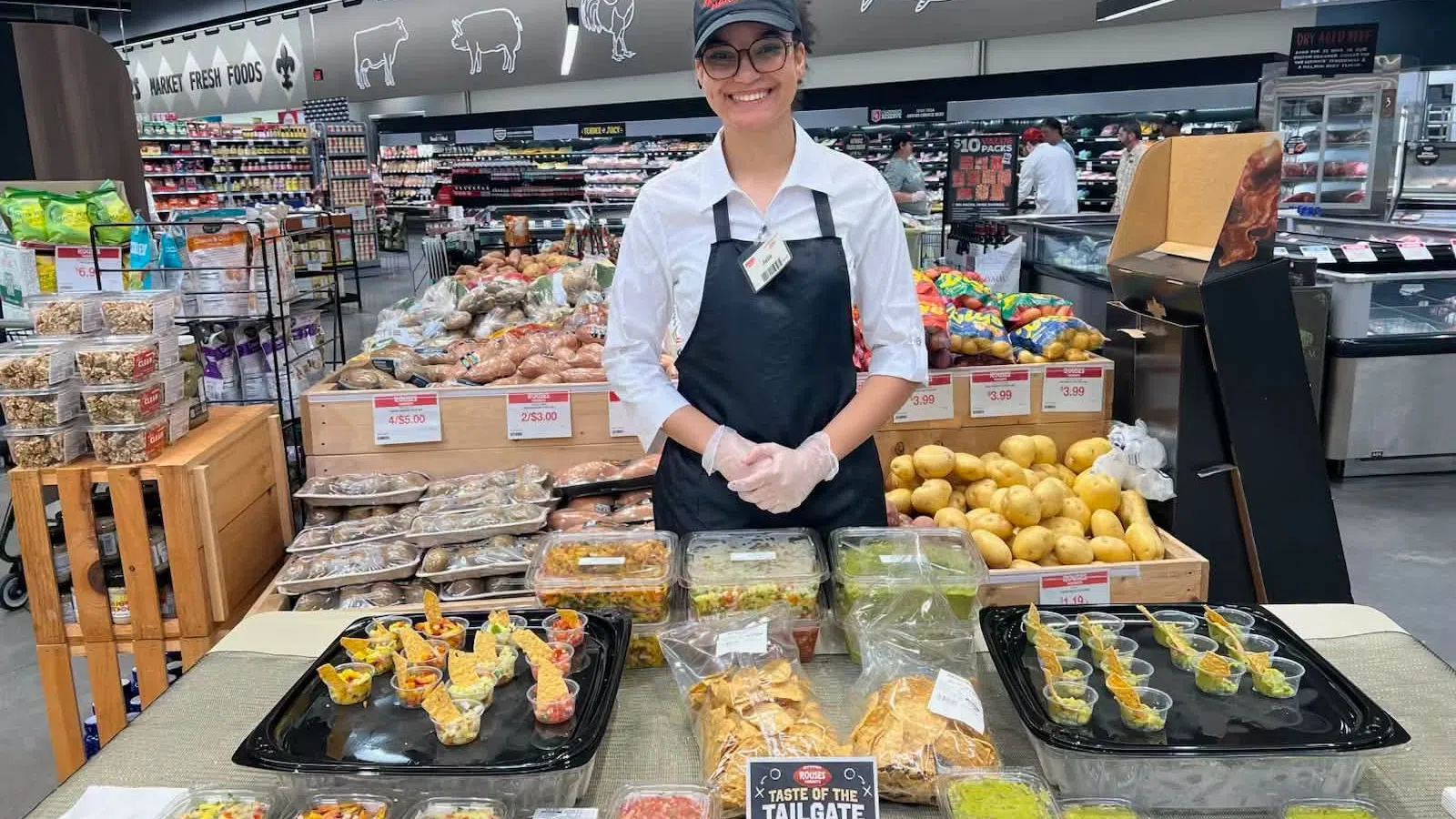 Smiling store employee presenting various homemade dips and salsas at a grocery store sampling table.