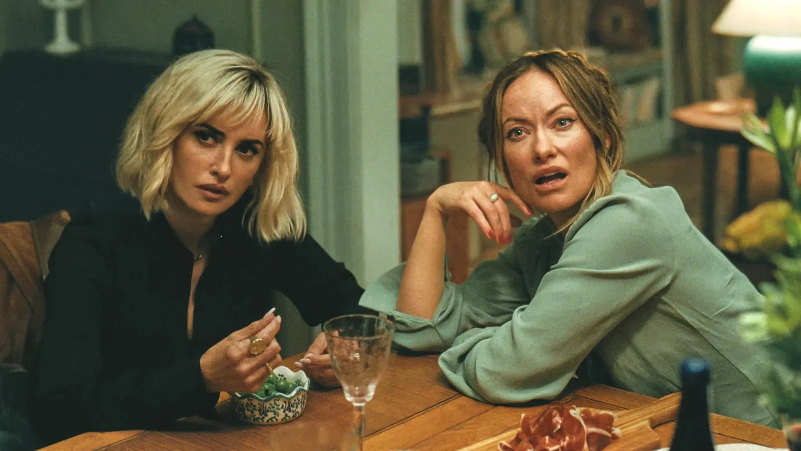 Two women with serious expressions sitting at a wooden table in a cozy, warmly lit room.