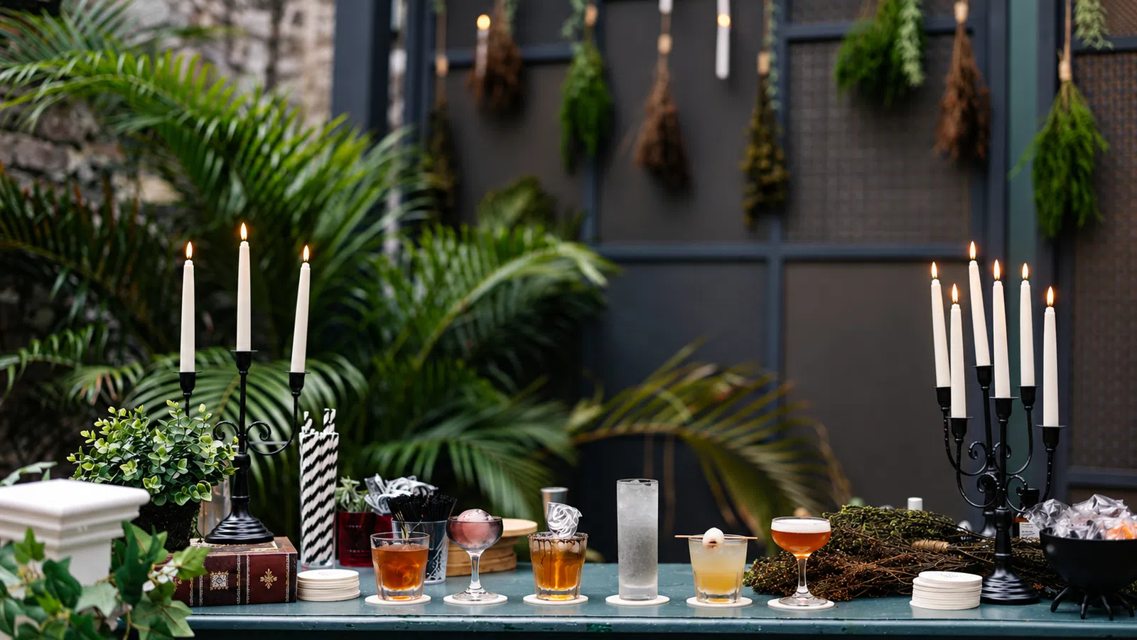 Outdoor bar setup with assorted cocktails, lit candles, greenery, and dried herbs hanging on a dark backdrop.