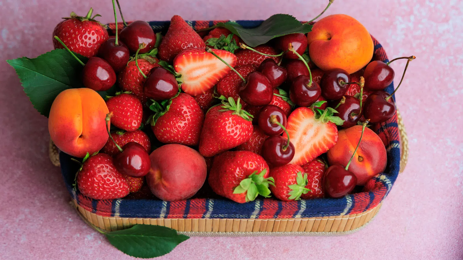 Basket filled with fresh strawberries, cherries, peaches, and apricots on a pink surface