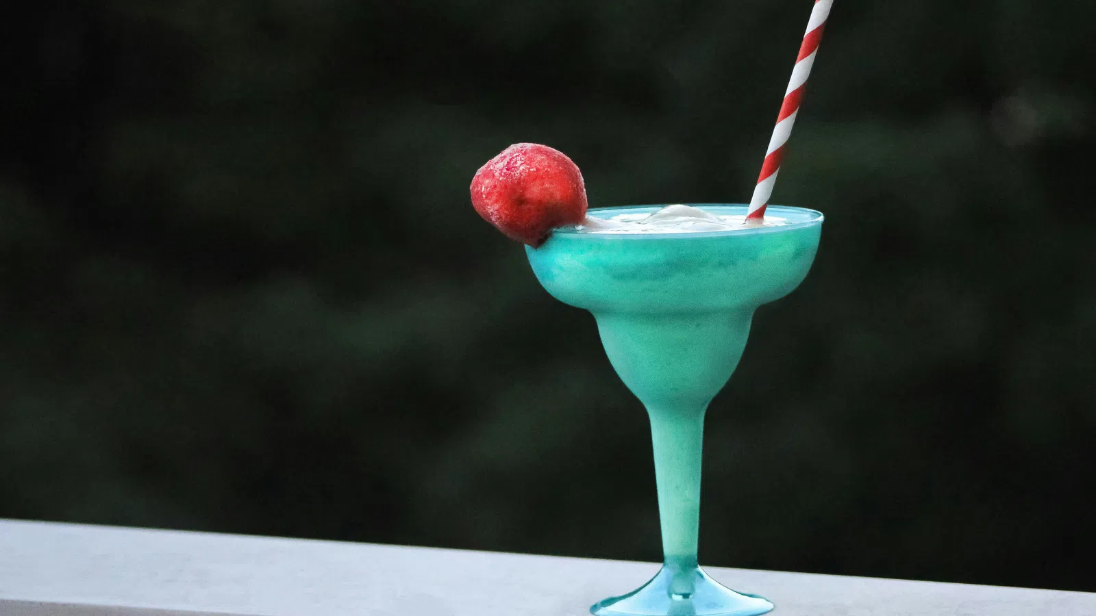 Blue tropical cocktail in a glass with ice, garnished with a red fruit and striped straw, on a white ledge.