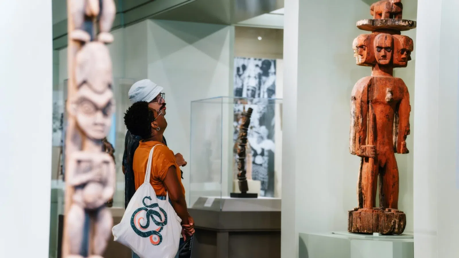 Visitors observe traditional wooden statues with carved faces at a museum exhibit focused on tribal art