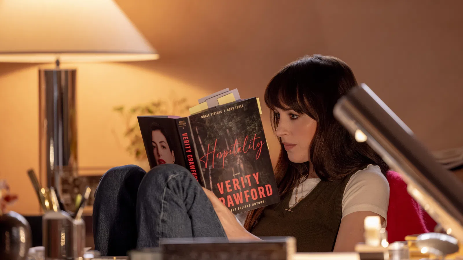Woman reading the book Hospitality by Verity Crawford in a cozy, warmly lit room with a table lamp.