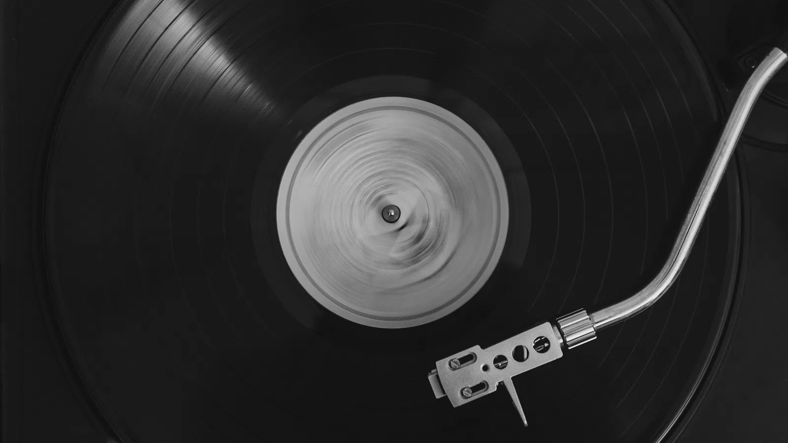 Black vinyl record spinning on a turntable with a silver tonearm in black and white photo.