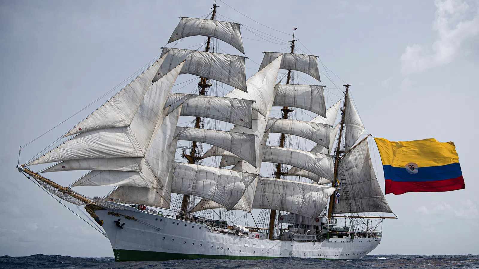 Large white sailing ship with multiple sails and a Colombian flag on the ocean under clear sky