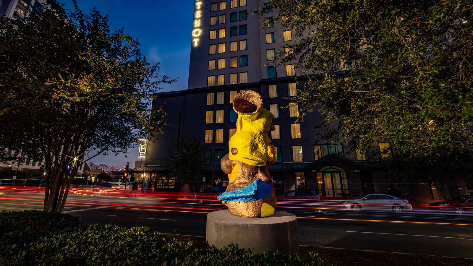 Yellow and blue abstract sculpture on a city street at night with building and light trails in background