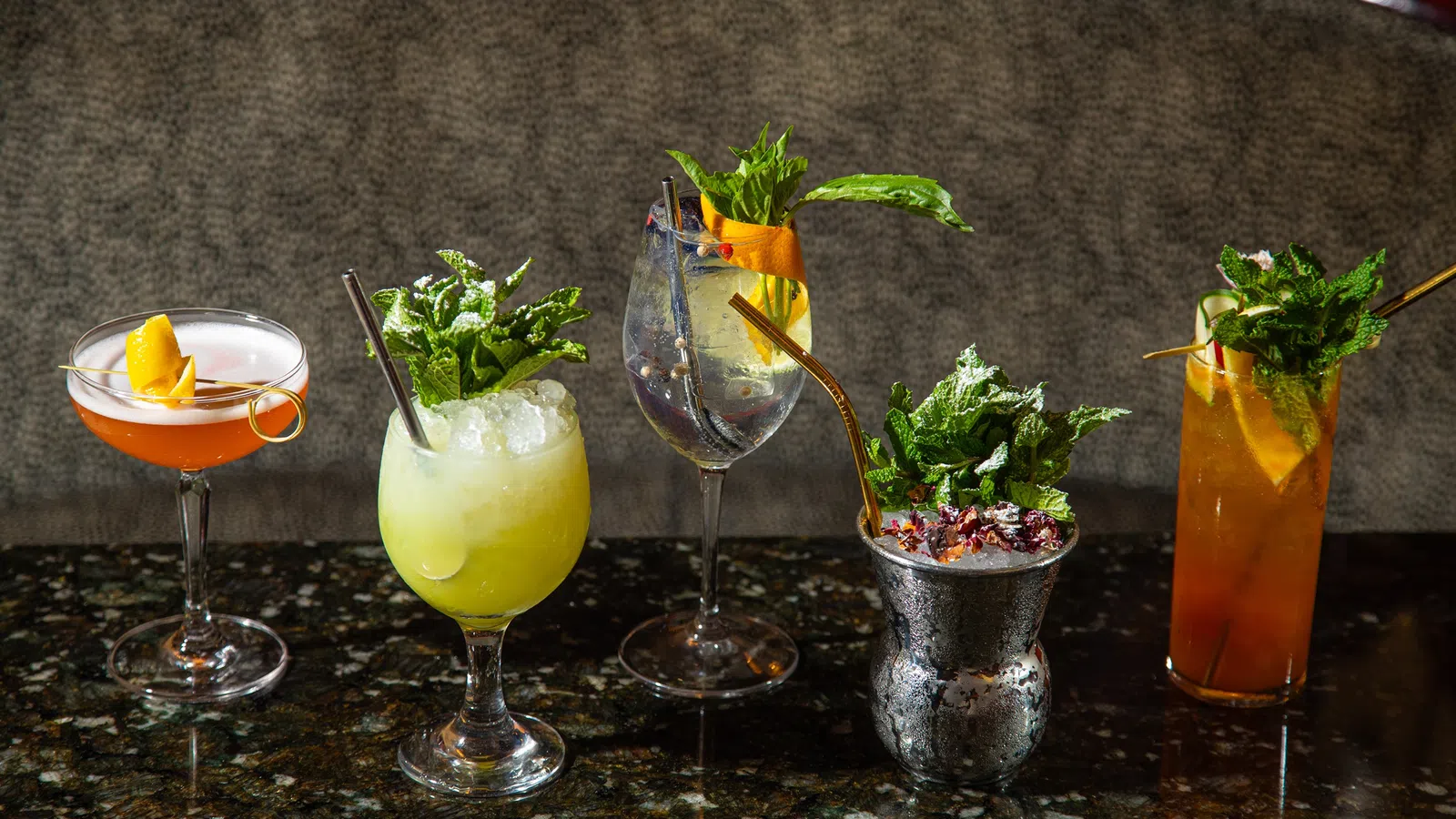 Five colorful cocktails with garnishes like mint, citrus, and berries on a dark granite bar counter.