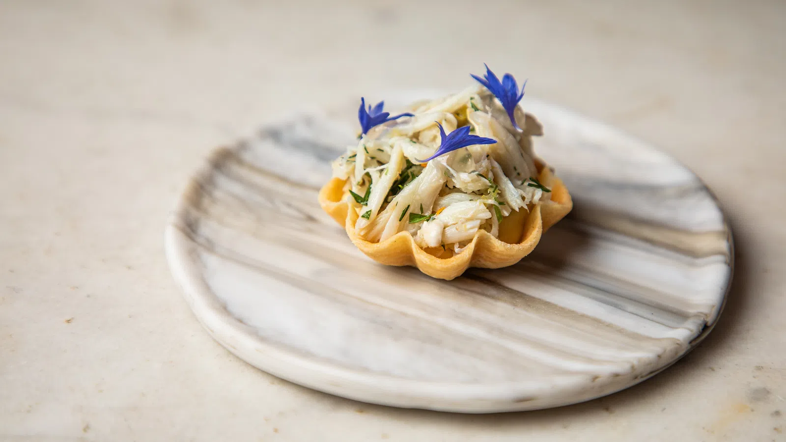 Gourmet tartlet filled with crab meat and garnished with blue edible flowers on a marble plate.