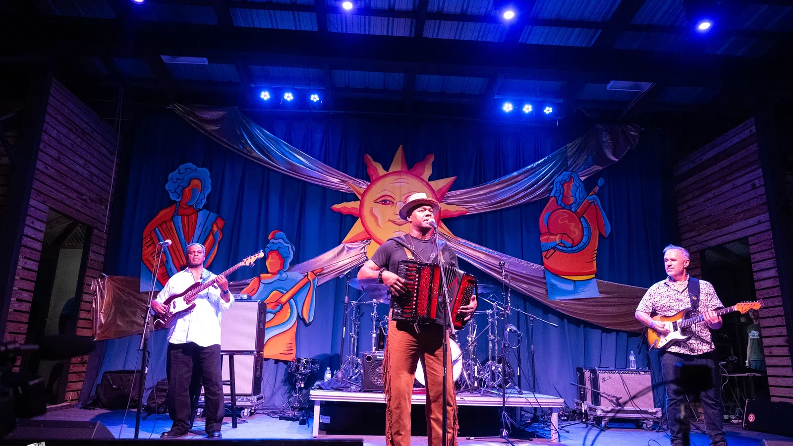 Live band performing on stage with accordion, guitar, and bass under bright lights and colorful sun backdrop