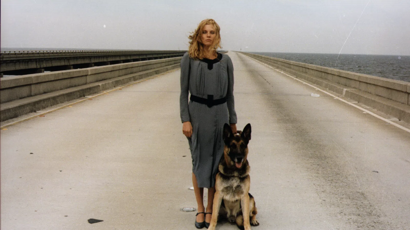 Woman in gray dress stands with German Shepherd on empty bridge over water under cloudy sky.