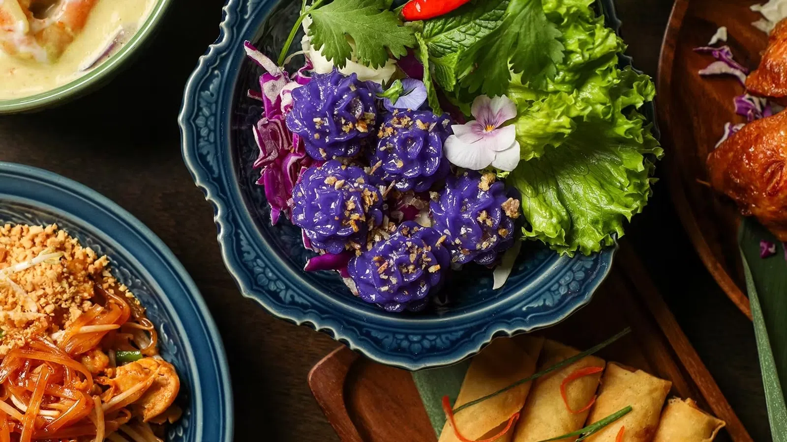 Colorful Thai dishes including purple flower-shaped dumplings, spring rolls, pad thai, and curry on ornate plates.