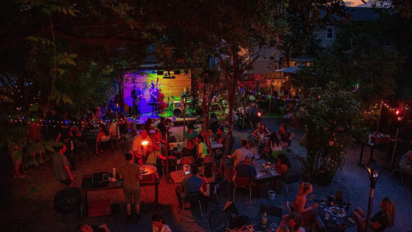 Outdoor evening concert with people seated at tables, colorful stage lights, and festive string lights among trees.