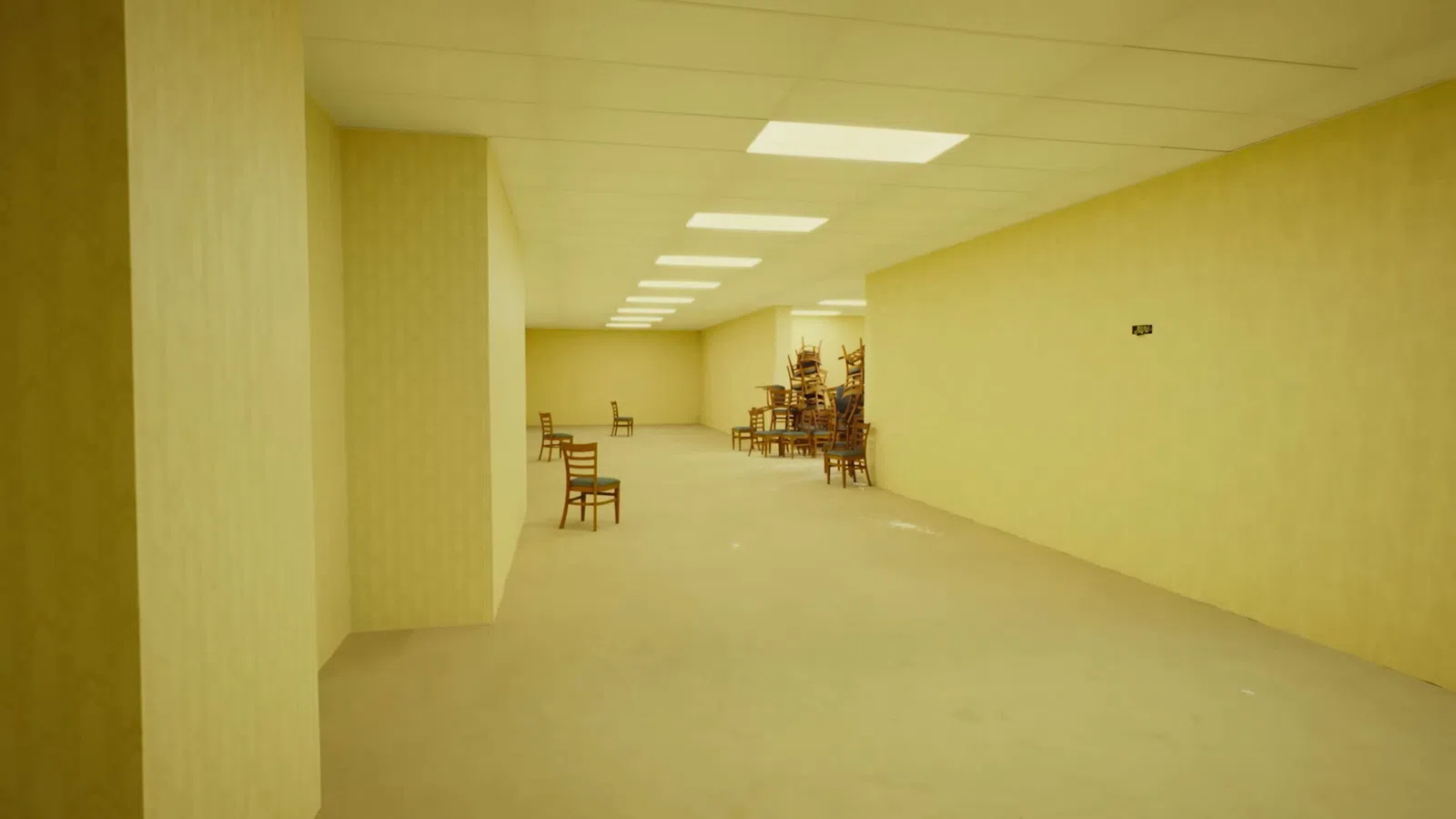 Empty beige room with chairs sparsely placed and a large stack of chairs in the corner under ceiling lights