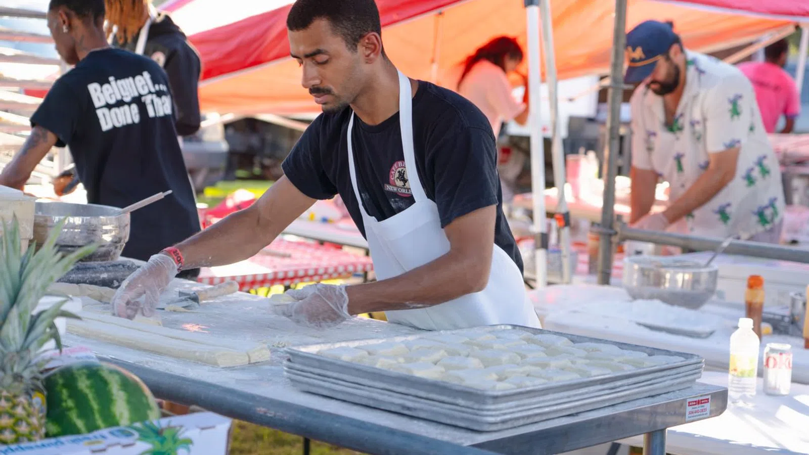 Man wearing apron preparing dough at outdoor food stall with trays of dough and tents in background