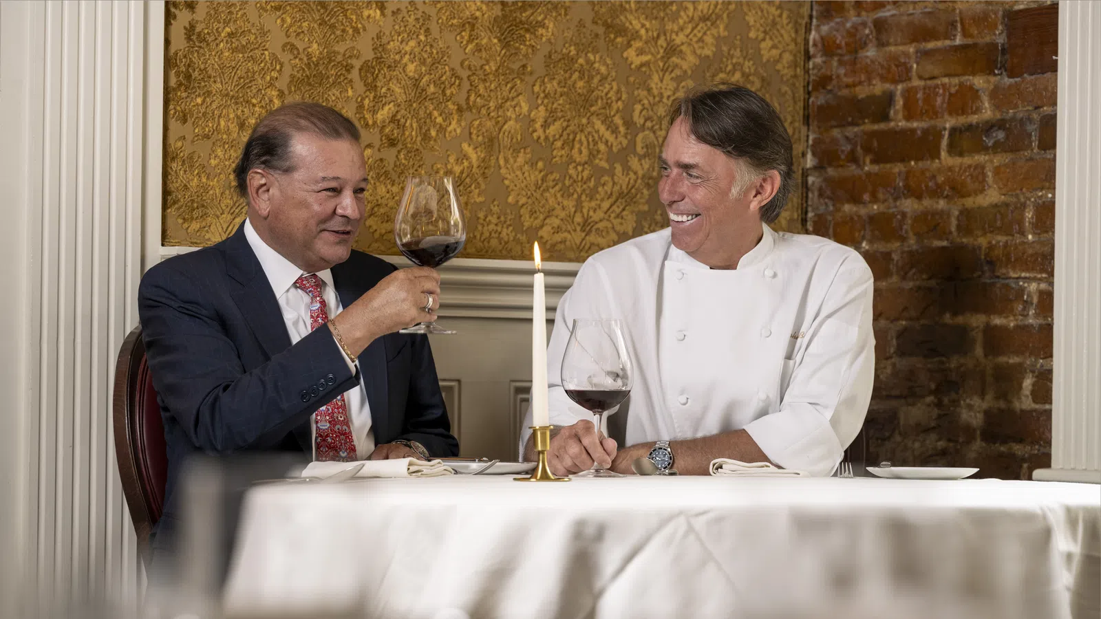 Two men smiling and toasting with red wine in an elegant dining room with gold wallpaper and exposed brick.