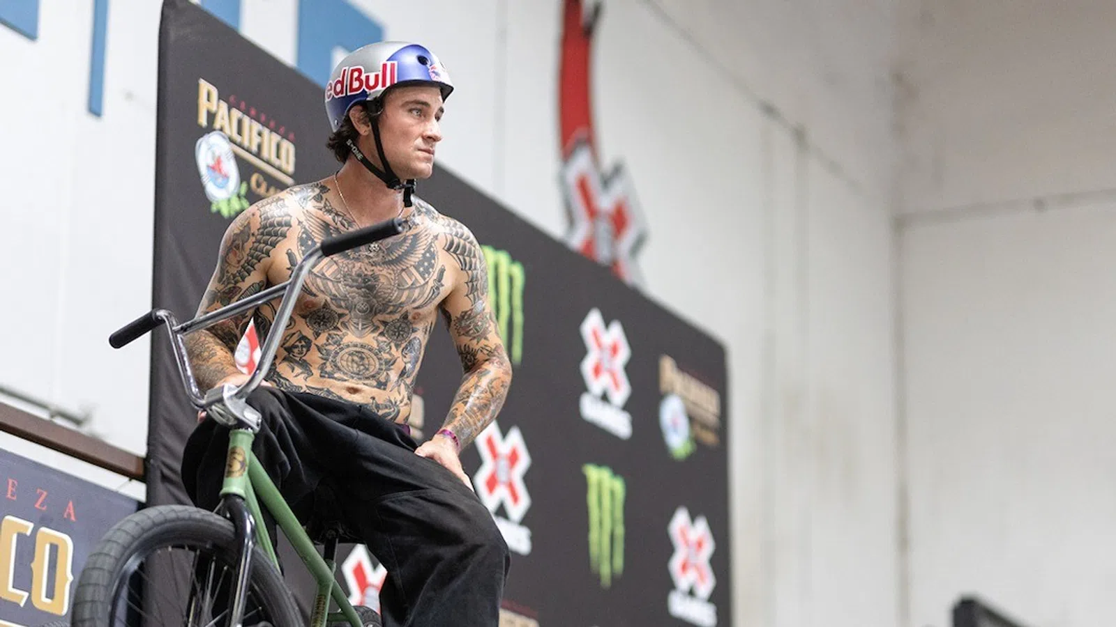 Tattooed BMX rider wearing Red Bull helmet posing with bike at indoor X Games skatepark event.