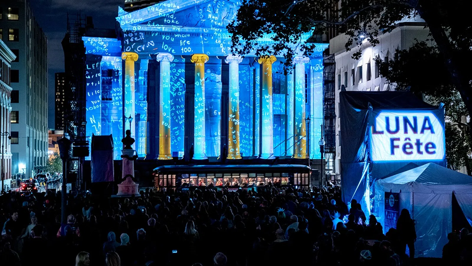 Crowd at night watching blue and gold light projections on a classical building during Luna Fête festival.