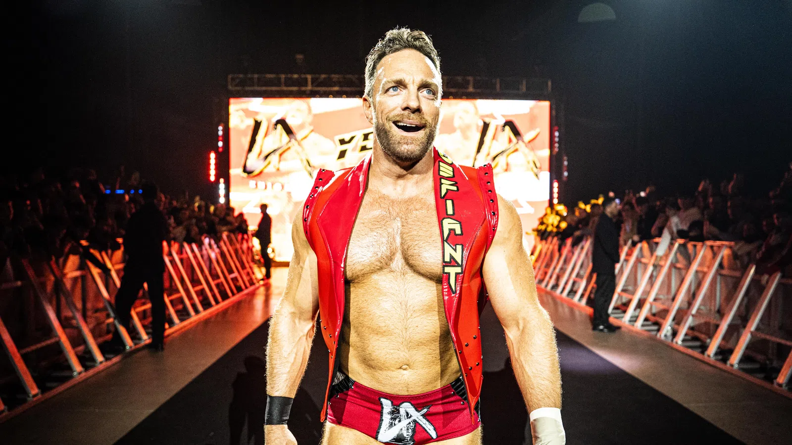 Muscular male wrestler in red trunks and vest walking confidently toward ring with audience and lights behind him