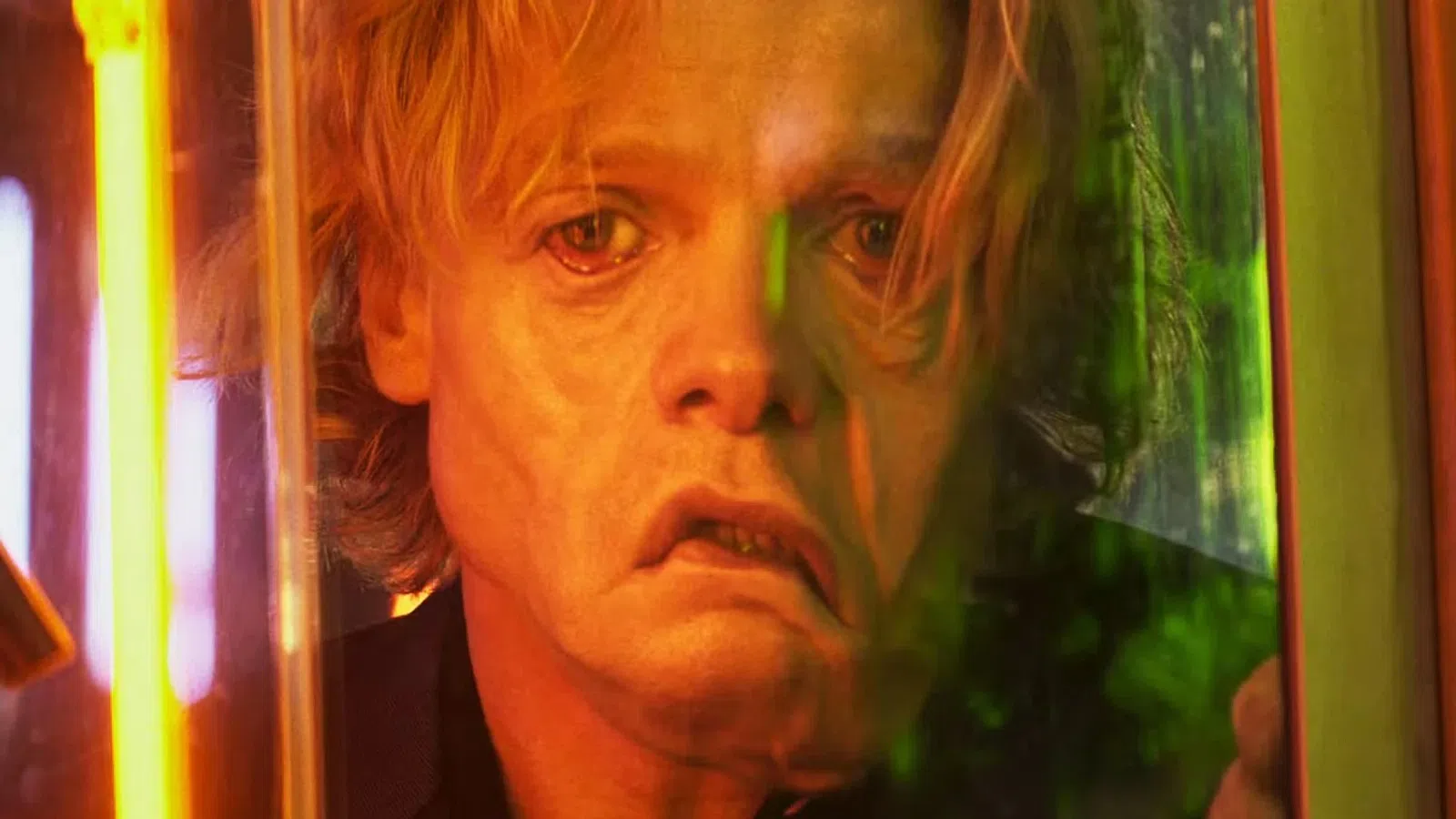 Close-up of a distressed man with blonde hair looking through a glass panel illuminated by orange and green light.