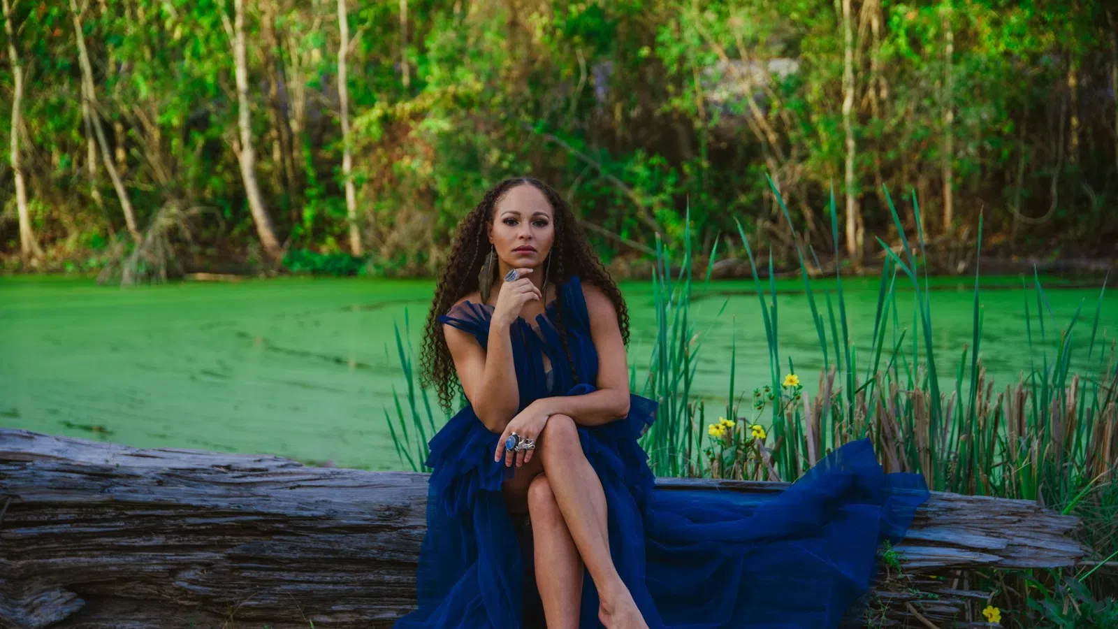 Woman in a flowing navy blue dress sitting barefoot on a log by a green pond surrounded by trees and plants.