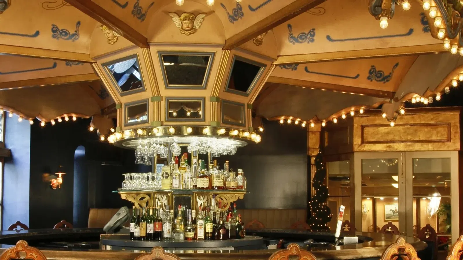 Elegant vintage bar interior with ornate ceiling, lights, central liquor display, and wooden chairs under warm lighting.