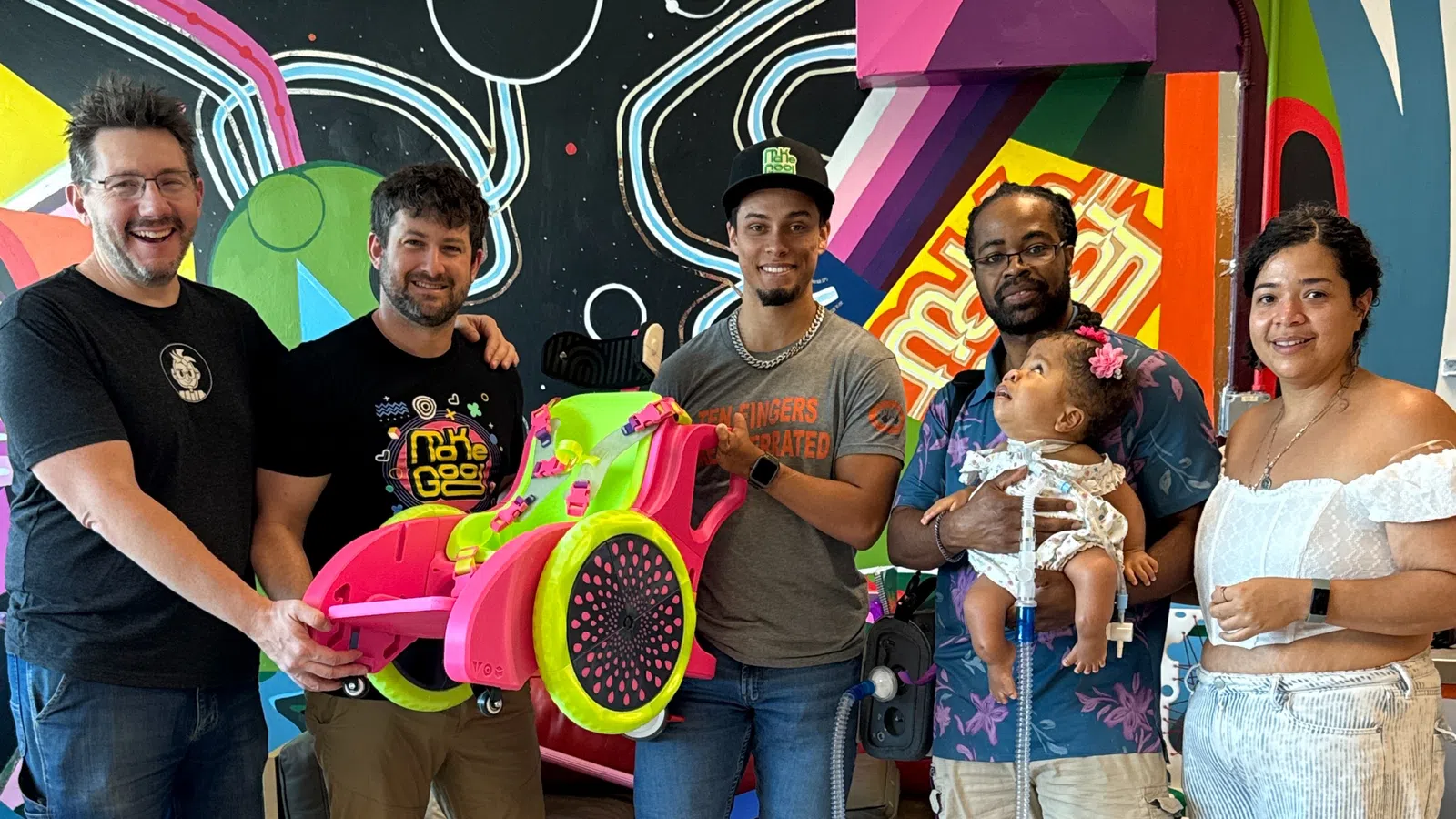 Group of five people smiling indoors with colorful wall art, holding a bright pink and green custom wheelchair.
