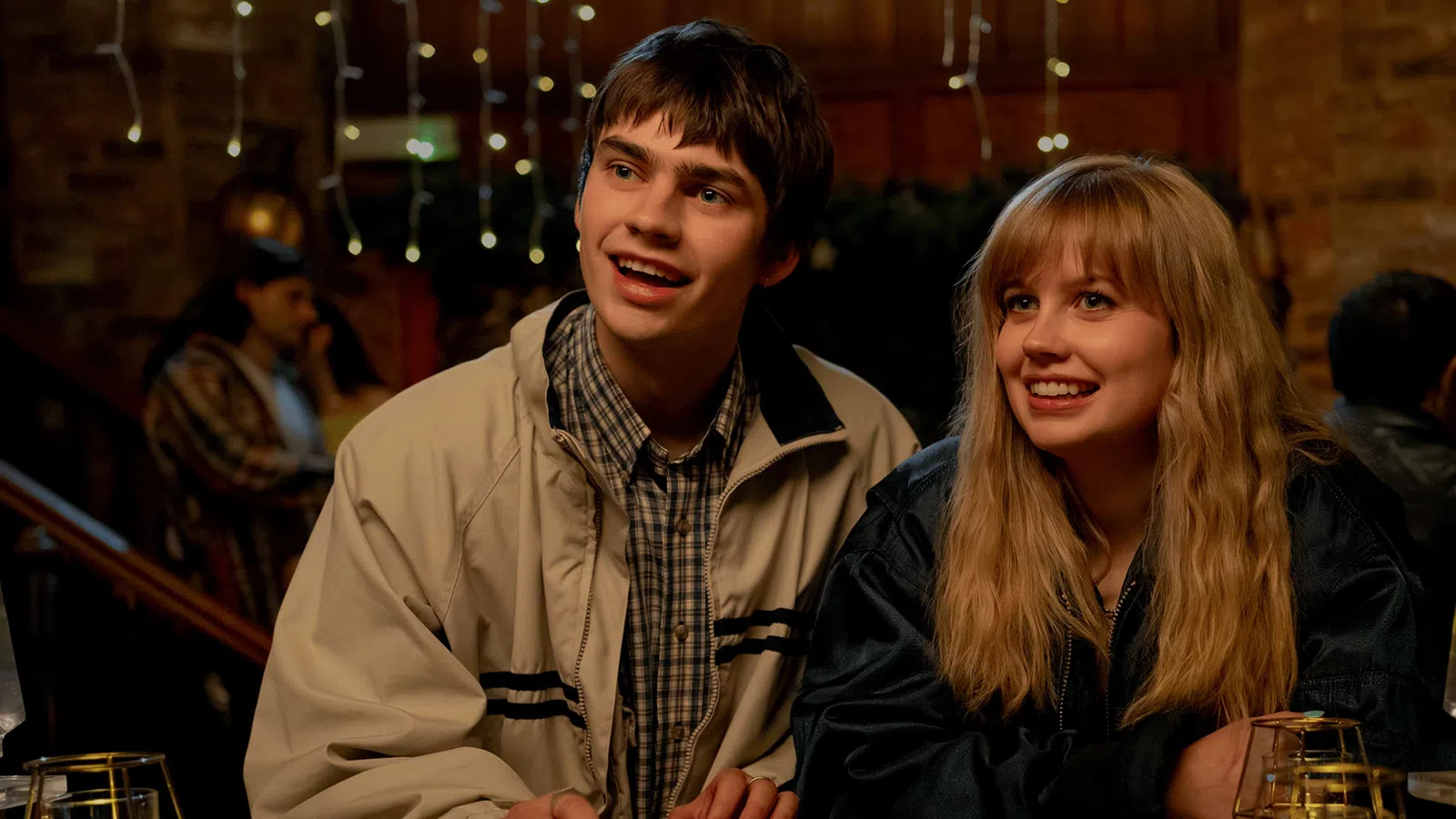 Teen boy and girl smiling and sitting together in a warmly lit indoor setting with people in the background