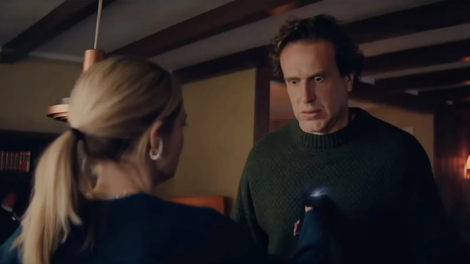 A man in a green sweater looks surprised as a woman with a ponytail holds out her hand indoors.