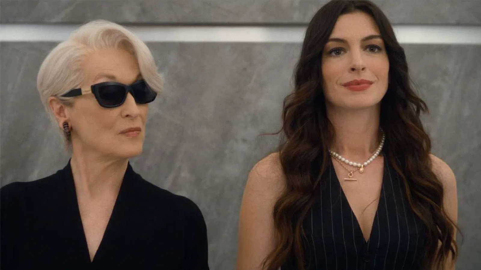 Two women in elegant attire, one with short white hair and sunglasses, the other with long dark hair and pearls.