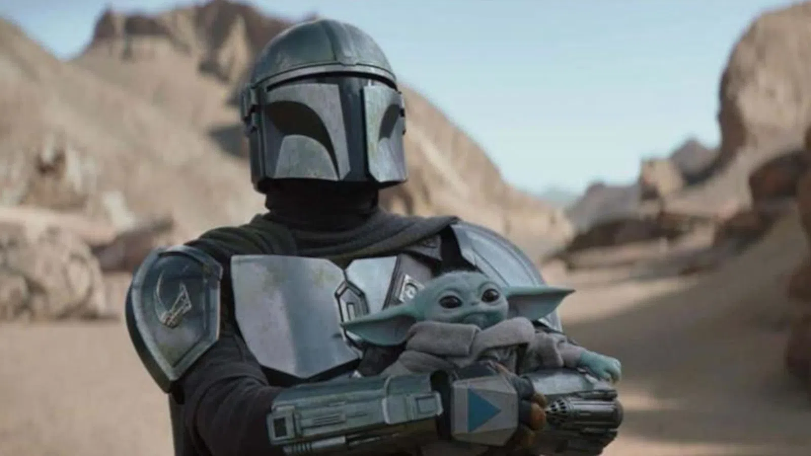 Armored Mandalorian holding Baby Yoda in desert landscape with rocky cliffs in background under clear sky