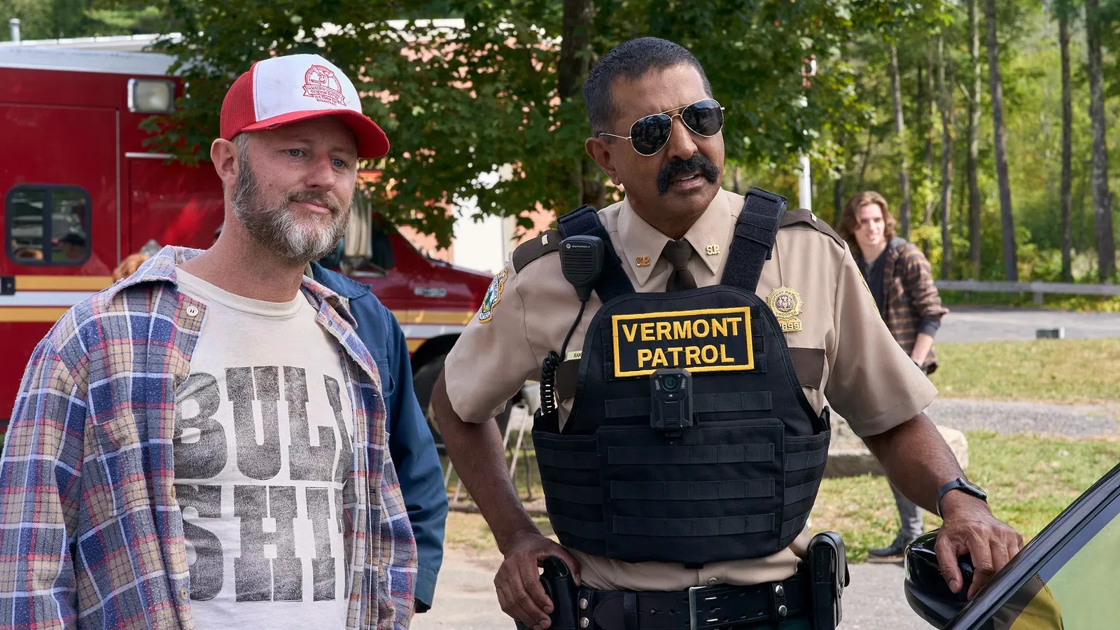 Vermont patrol officer in uniform speaking with a casually dressed man outdoors near an emergency vehicle.