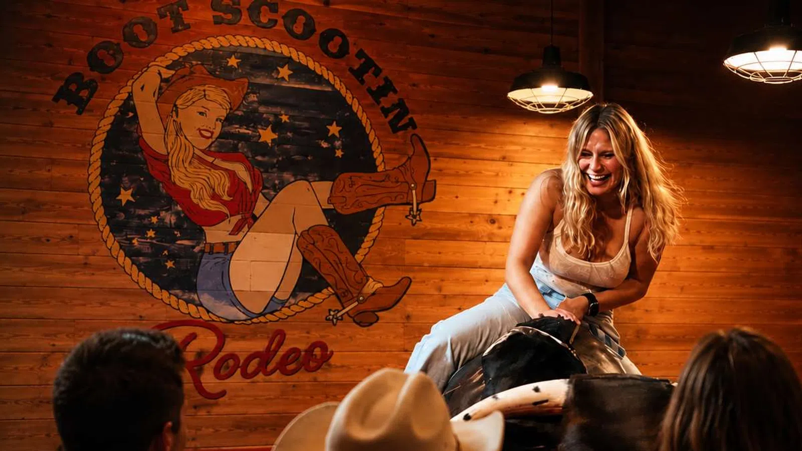 Young woman smiling while riding a mechanical bull in a wooden rodeo-themed bar with audience watching.