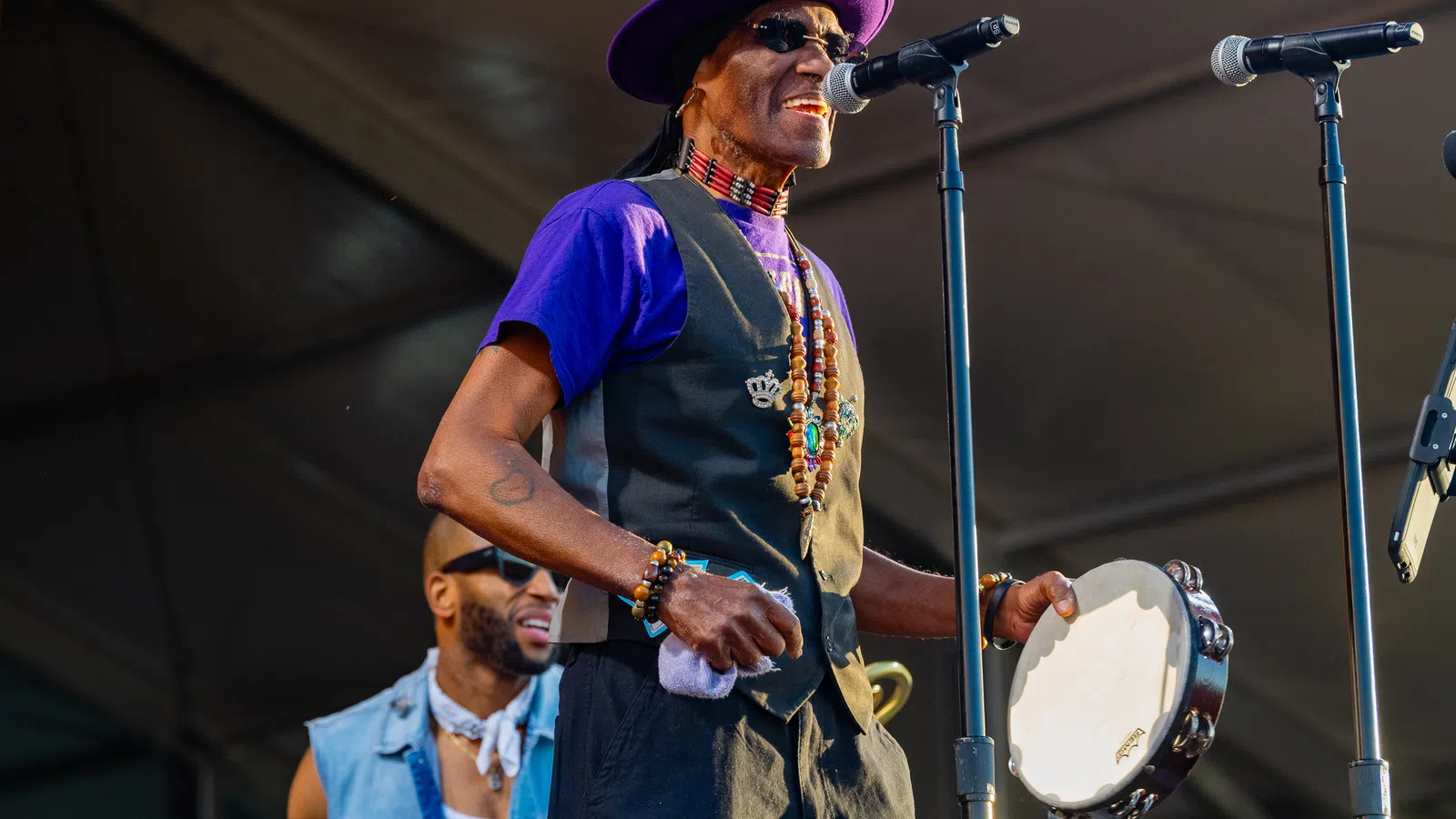 Musician in purple hat and vest singing into microphone while playing tambourine on stage at outdoor event.