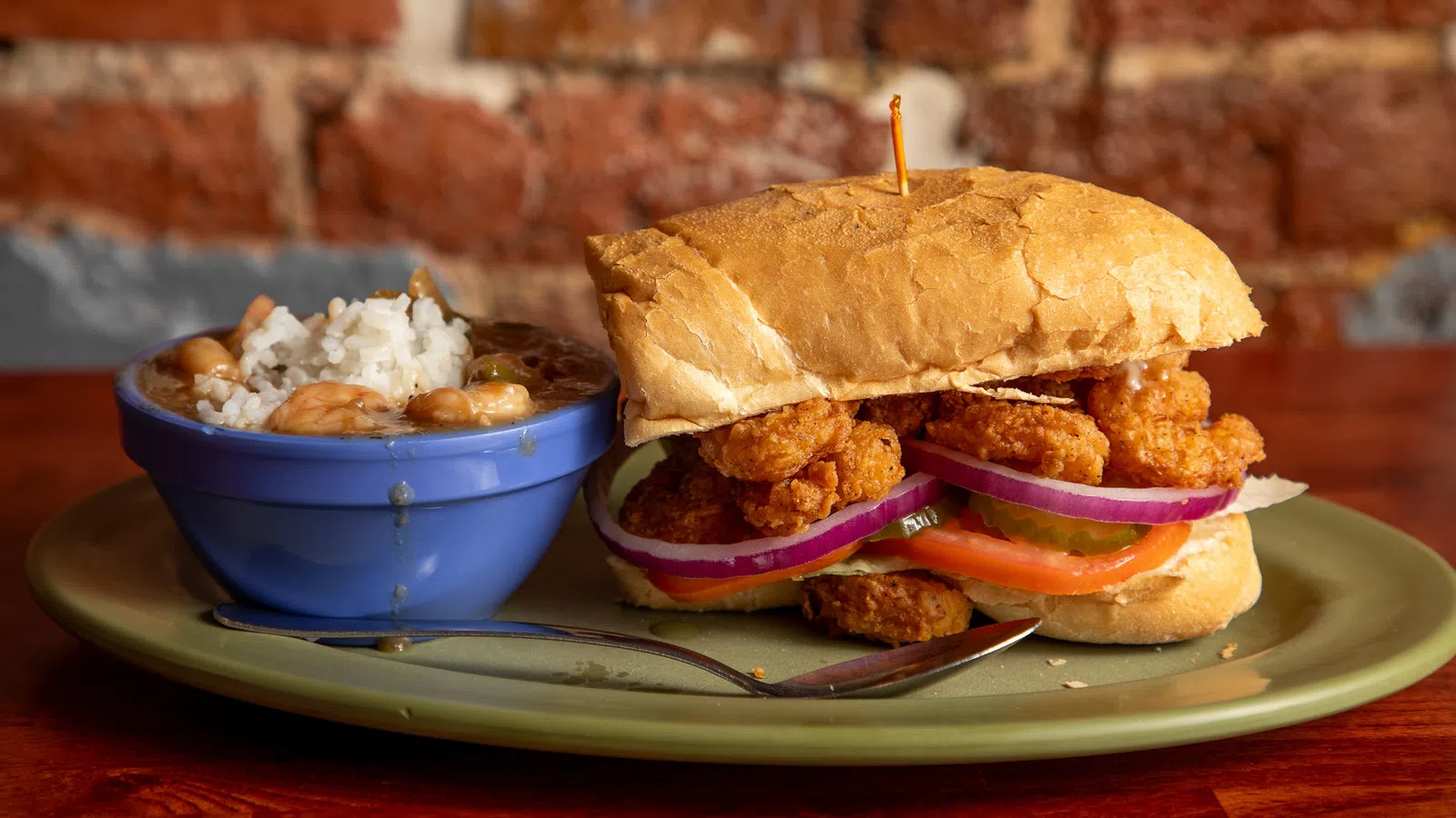 Crispy fried shrimp po' boy sandwich with lettuce, tomato, onion, served with a side of rice and beans in a blue bowl.