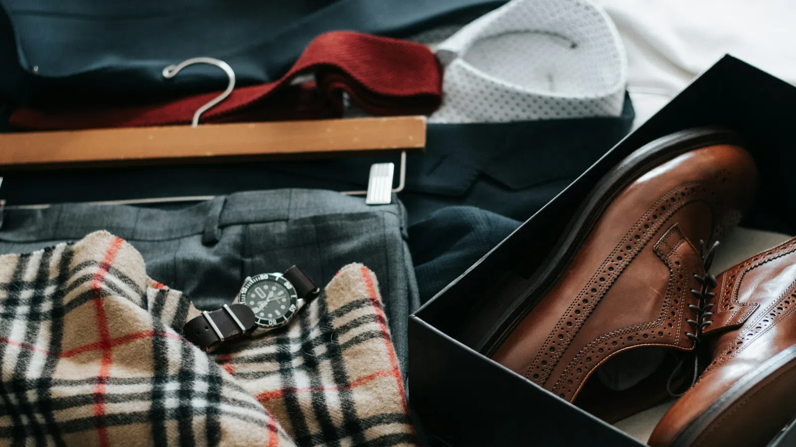 Flat lay of men's brown dress shoes in a box, plaid scarf, gray trousers, watch, and a white shirt on a dark suit jacket.