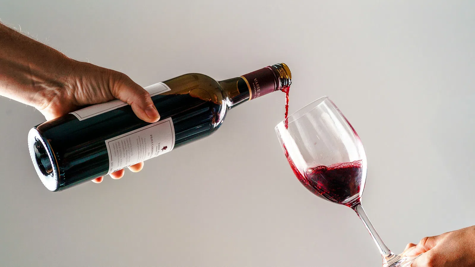 Red wine being poured from a bottle into a glass held by two hands against a plain background.