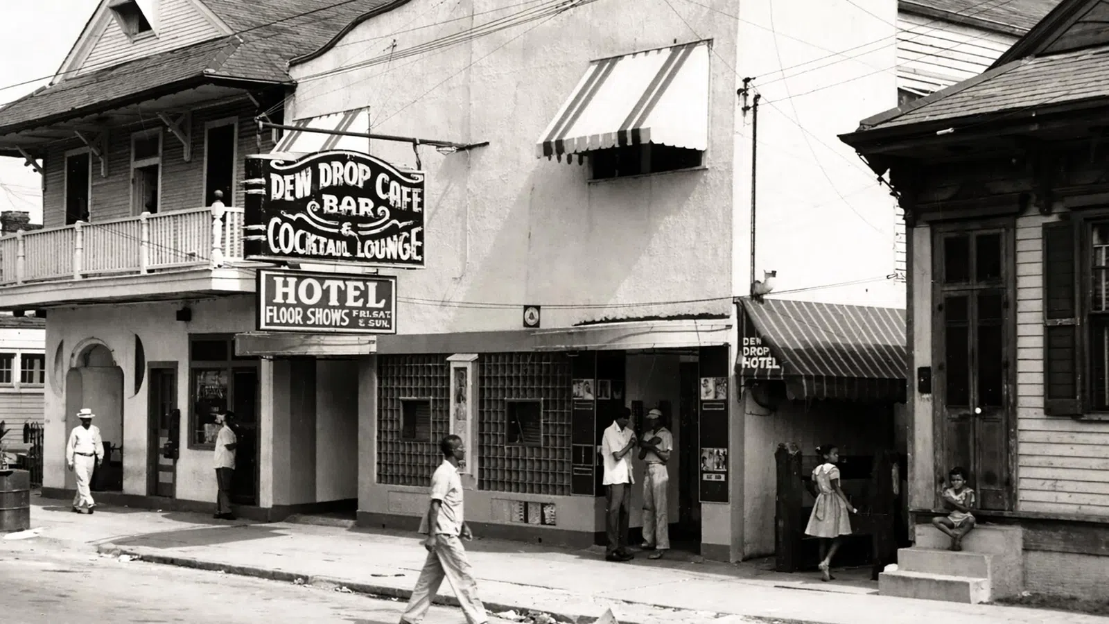 Black and white street scene with Dew Drop Cafe bar, cocktail lounge, hotel, and people walking and standing outside.