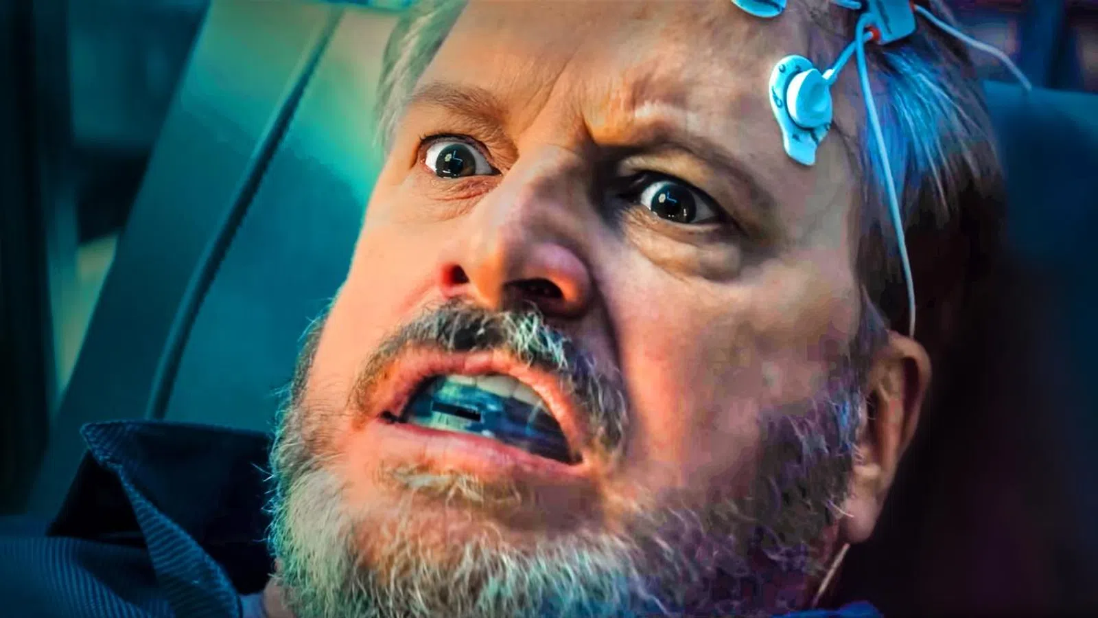 Man with electrodes attached to his head wearing a mouthguard, showing a fearful expression in a dark setting.