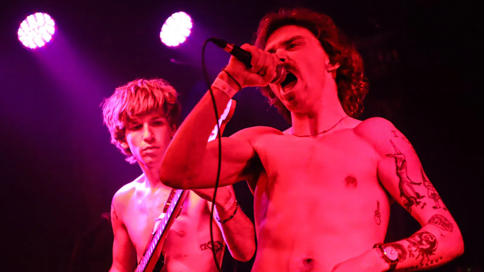 Two shirtless male musicians perform on stage under pink and purple lights, one singing and one playing guitar.