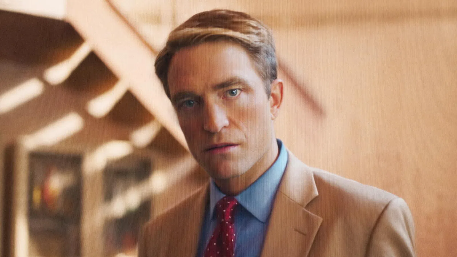 Man in a tan suit and red polka dot tie staring directly at the camera with a blurred warm background.